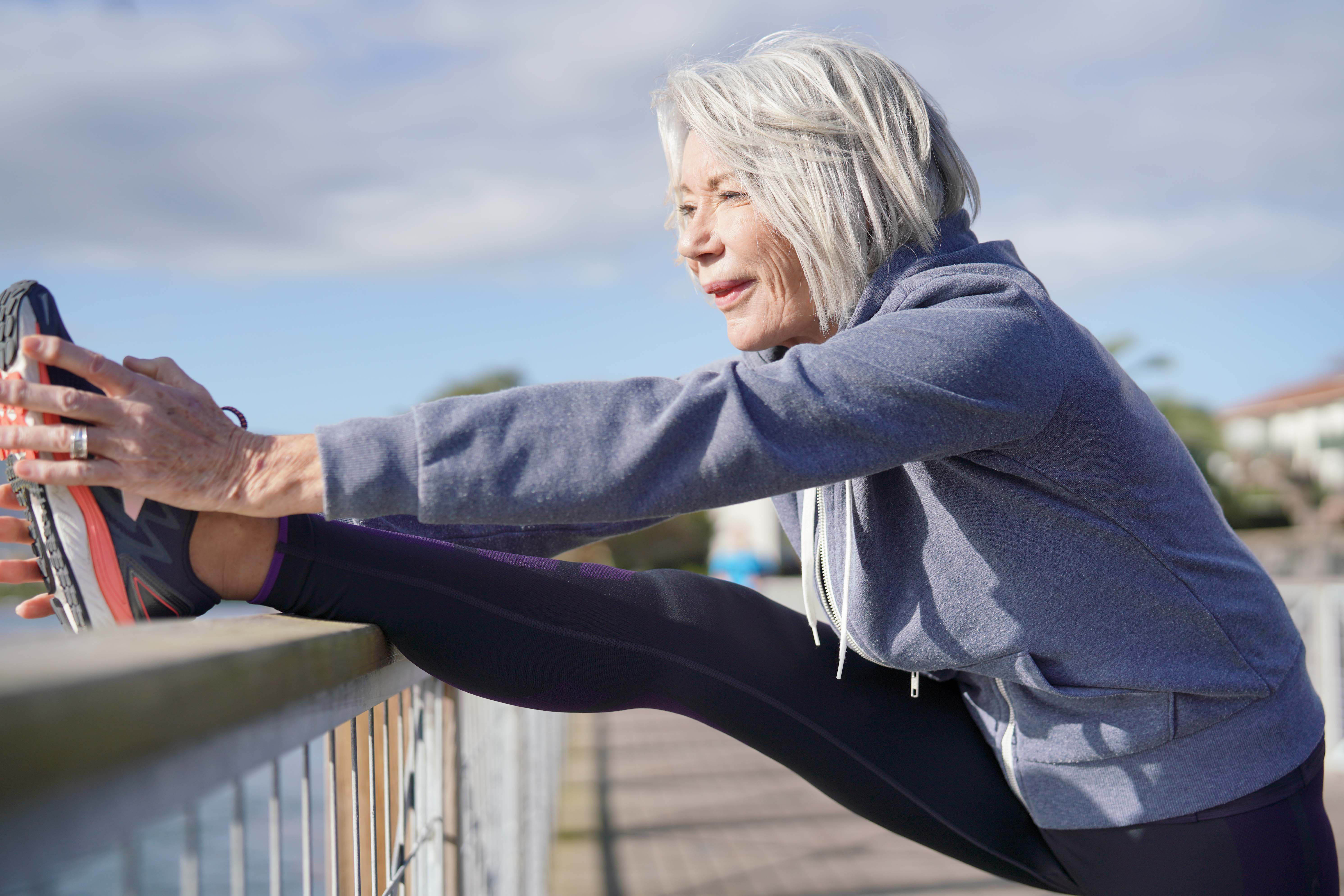 Older woman stretching her leg on a railing near the water Older woman stretching her leg on a railing near the water